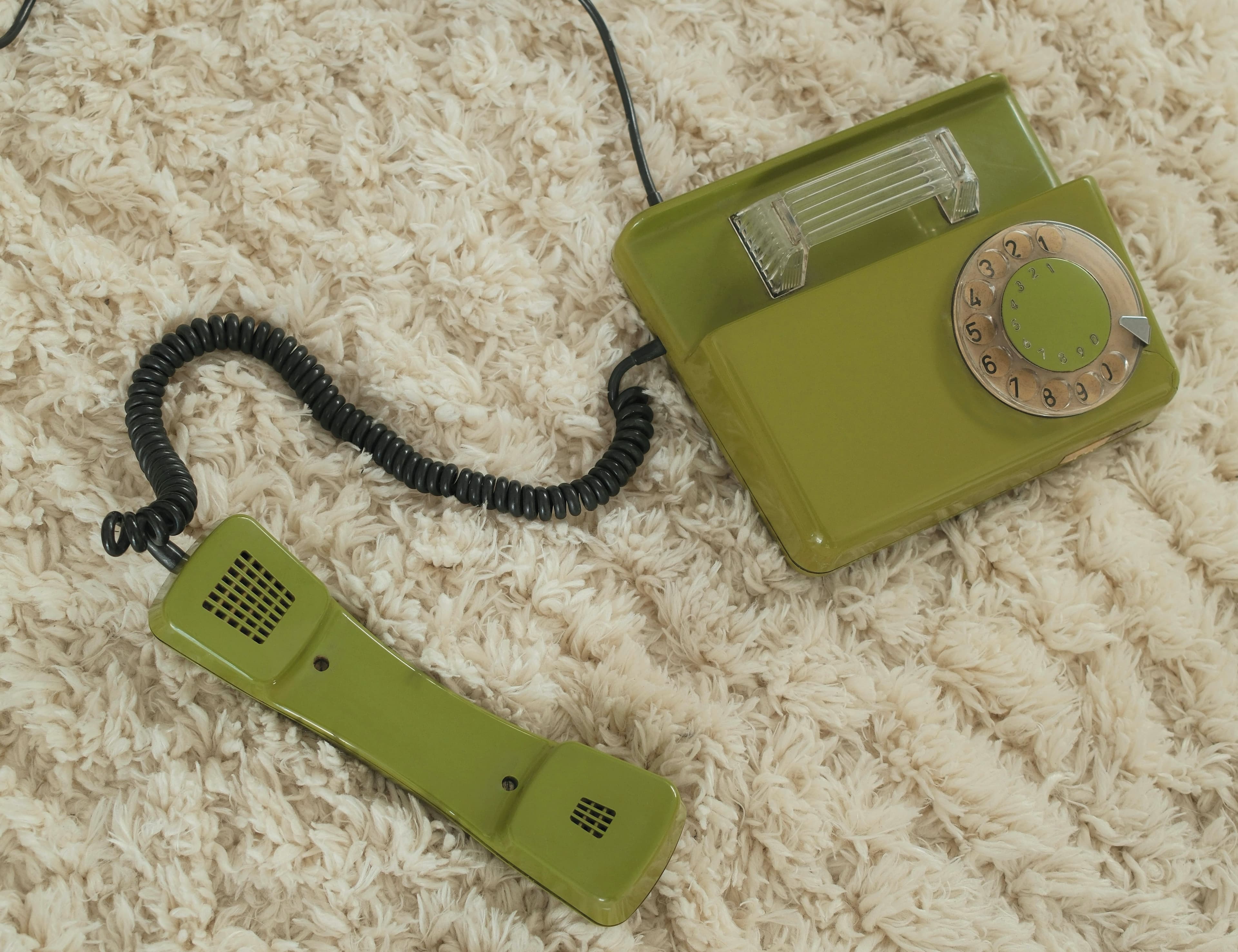 Green rotary phone with the handset off the hook, resting on a beige shag carpet.