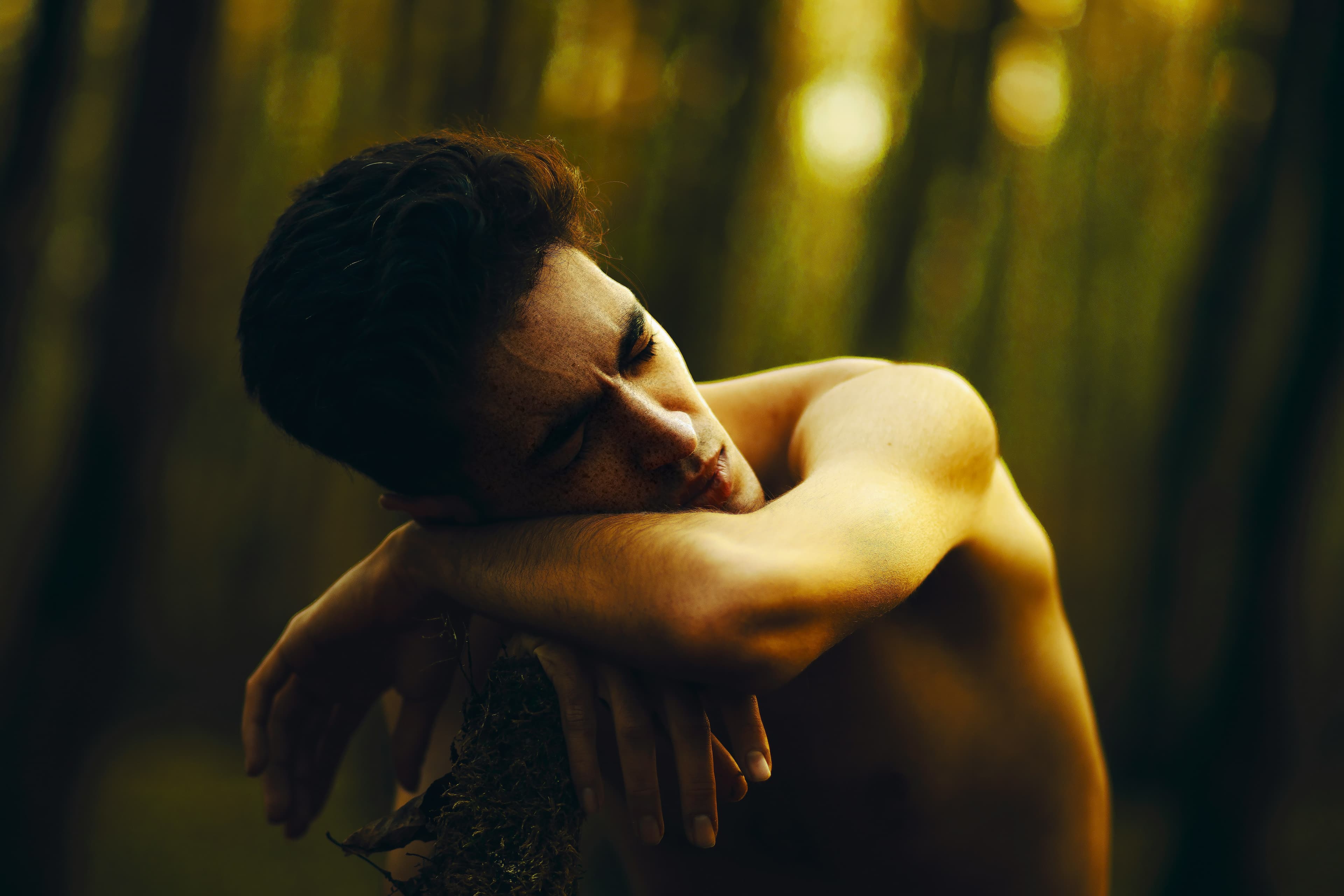 Shirtless person with short dark hair leans on a tree branch, eyes closed, in a sunlit forest setting.