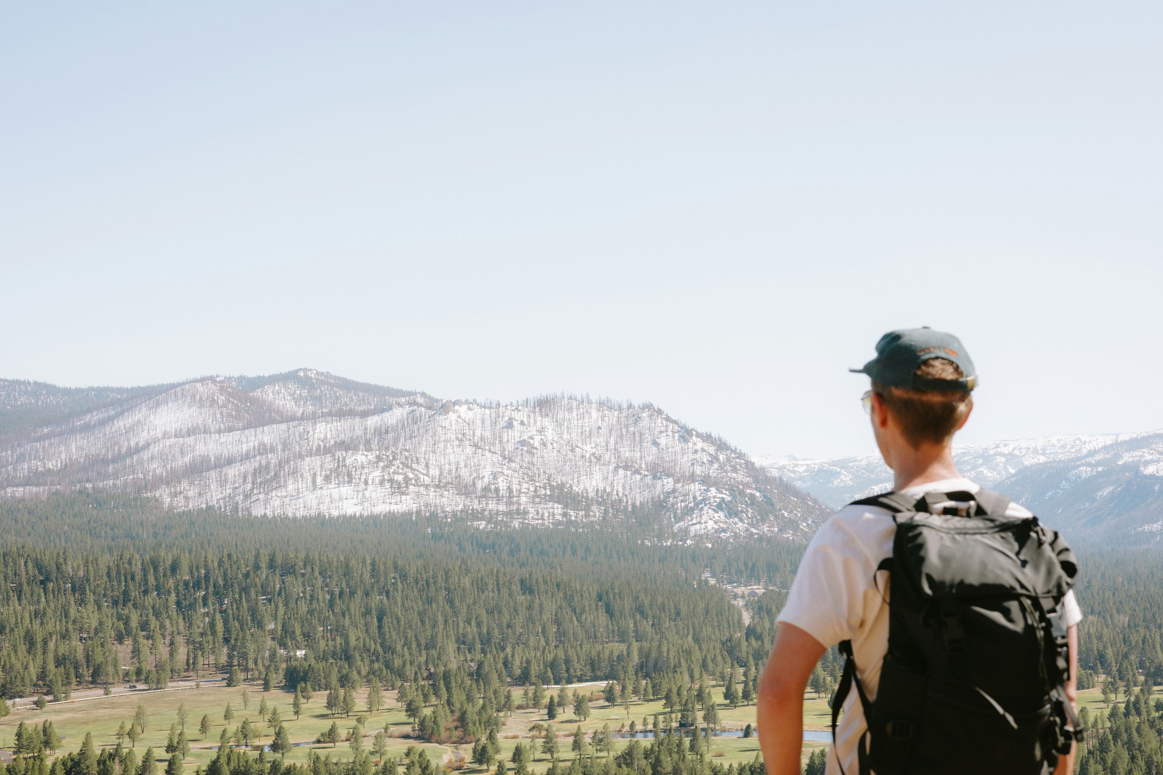 Person with a backpack and cap overlooks a vast forested valley and distant snow-capped mountains under a clear blue sky.